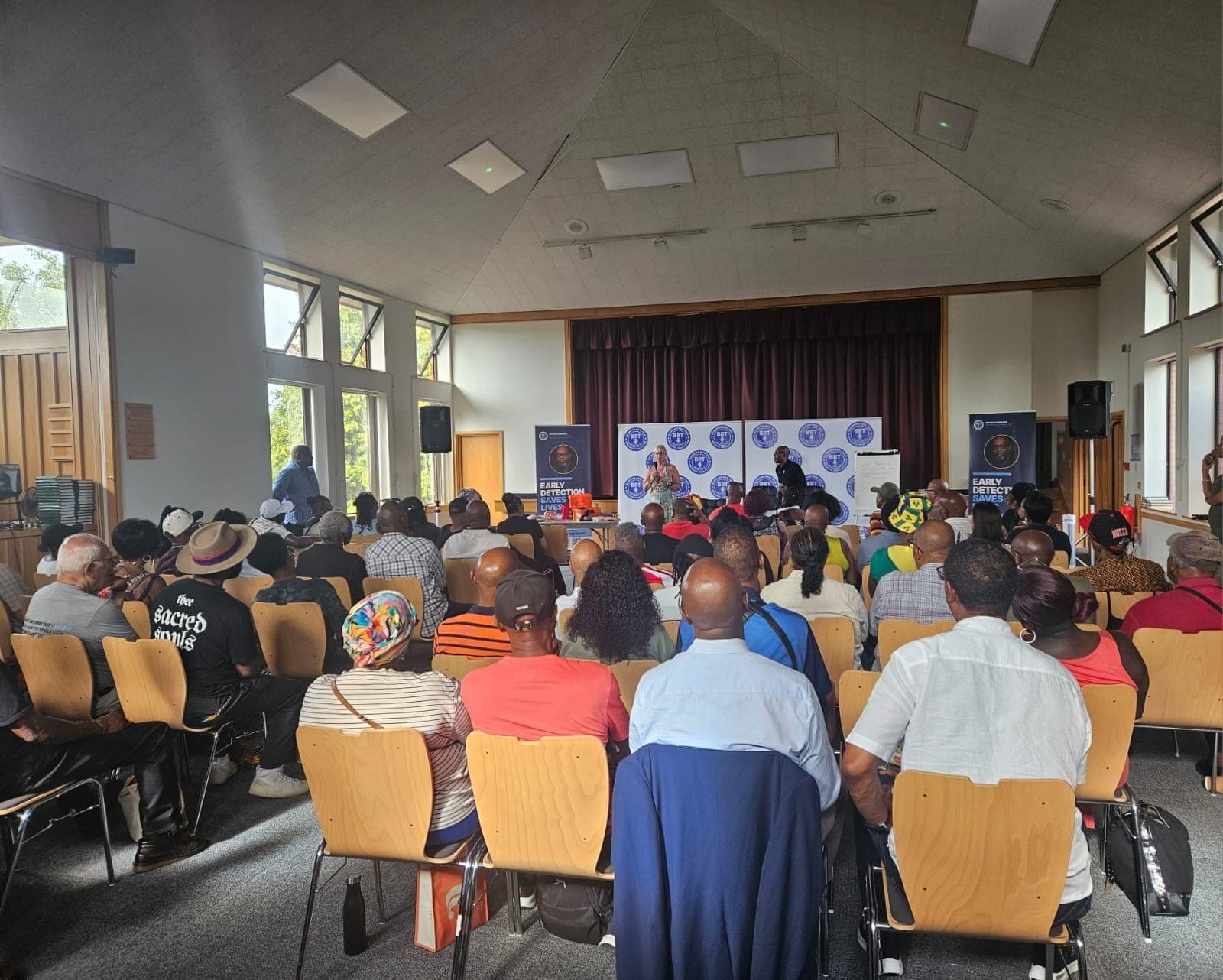 The full room photographed from the back, around sixty people seated and listening.